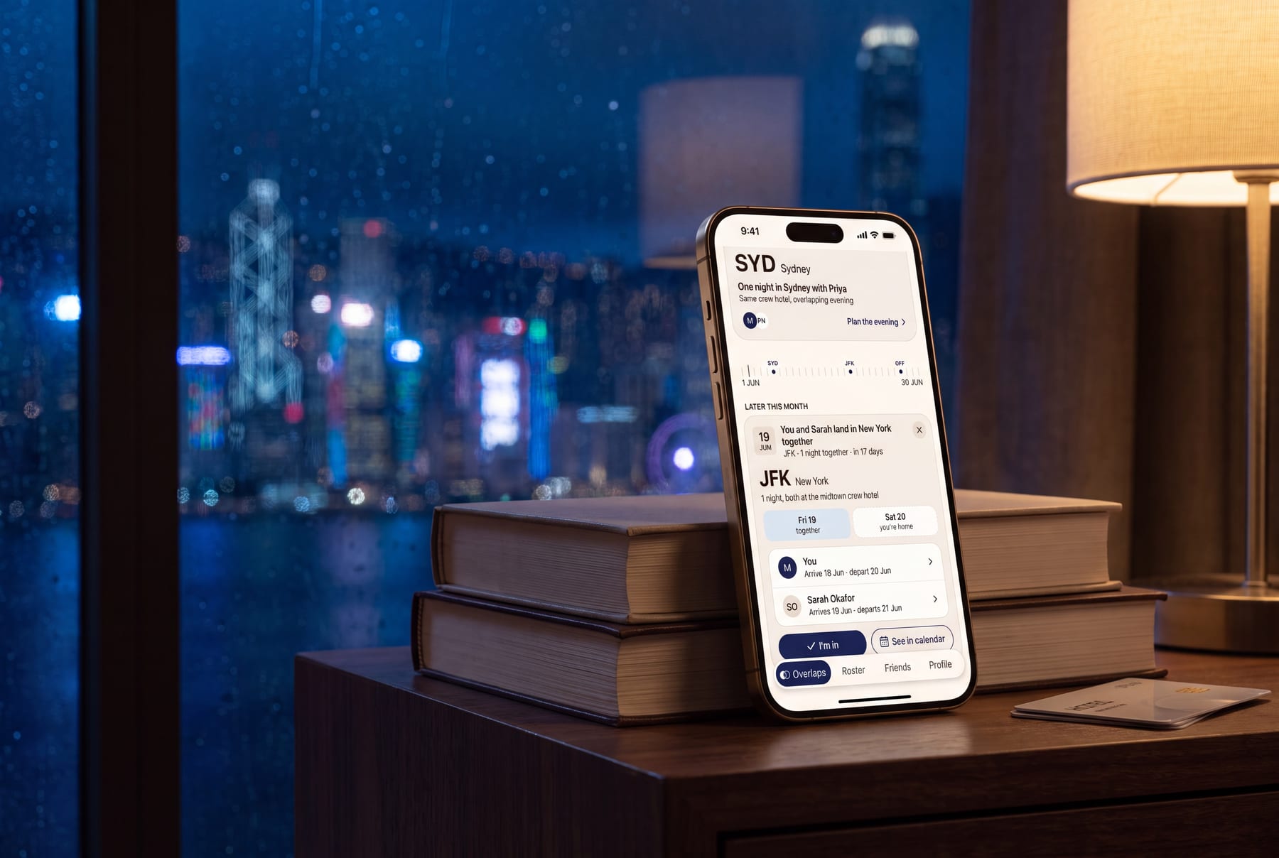 The Overlap on a hotel nightstand at night — an expanded New York layover card, with the Hong Kong skyline soft in the window